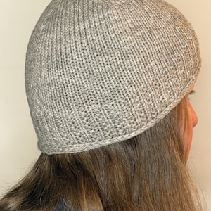 Könnte beinhalten: Eine hellgraue Strickmütze mit vertikaler Rippenstruktur über die gesamte Kappe und einem glatten, gerollten Rand an der Basis. Die Mütze ist in einem gleichmäßigen, feinmaschigen Strickmuster aus neutralem, meliertem grauen Garn gefertigt.
