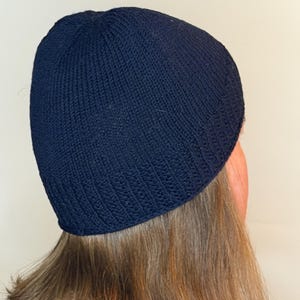 May include: A solid navy blue knit beanie cap featuring a ribbed texture along the bottom edge and a smooth stockinette stitch pattern on the crown. The hat is constructed from a dark blue yarn and fits snugly around the head with a simple design.
