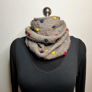 Puede incluir: Una bufanda infinita de punto color marrón claro con pequeños puntos en relieve de colores amarillo, rojo y azul. La bufanda está drapeada en forma circular y superpuesta, mostrando una textura de hilo suave en toda la prenda.