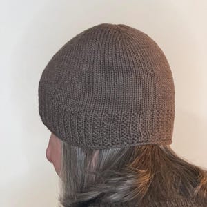 Könnte beinhalten: Eine braune Strickmütze. Die Mütze ist einfarbig und scheint aus einem weichen, warmen Material zu bestehen. Die Mütze ist ein eng anliegender Stil, perfekt für kaltes Wetter.