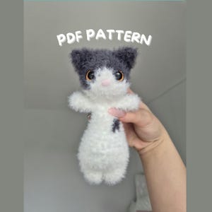 May include: A handmade, plush cat toy in white and grey, with large, expressive eyes. The cat has a small black spot on its chest. The text "PDF PATTERN" is displayed above the toy. The toy is held by a person's hand.