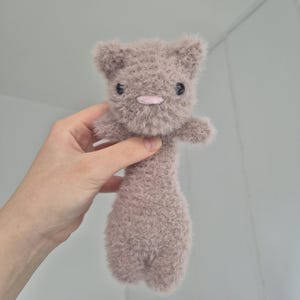 May include: A handmade, plush toy cat in a soft, muted mauve color. The cat has small, black button eyes, a pink nose, and a fluffy texture. The toy is held up by a hand.