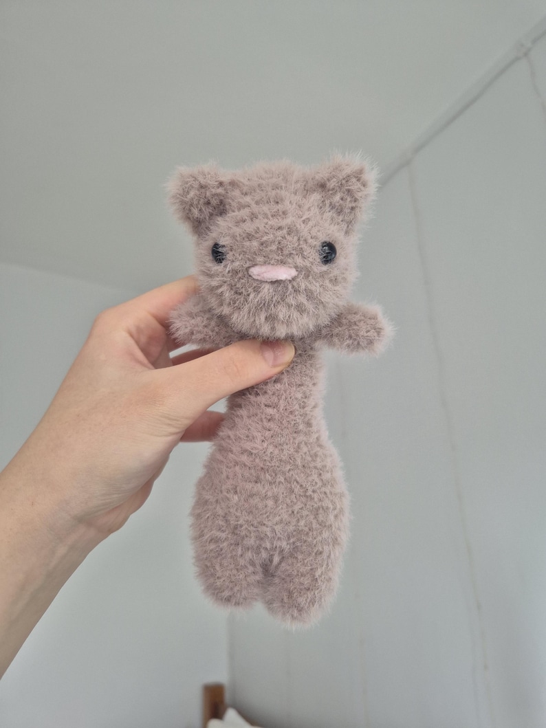 May include: A handmade, plush toy cat in a soft, muted mauve color. The cat has small, black button eyes, a pink nose, and a fluffy texture. The toy is held up by a hand.