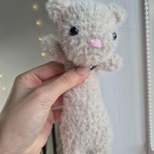 May include: A handmade plush toy cat in a light gray color. The cat has black eyes, a pink nose, and small arms. The plush toy is made of a soft, fuzzy material.