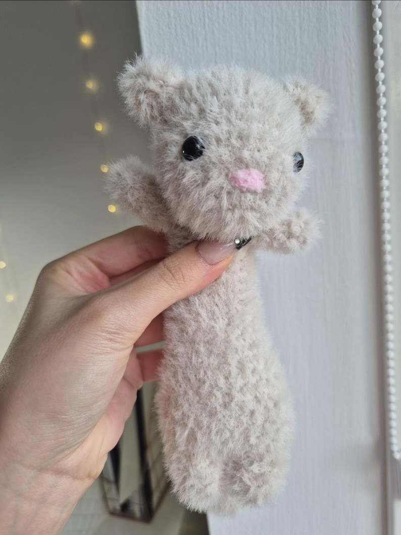 May include: A handmade plush toy cat in a light gray color. The cat has black eyes, a pink nose, and small arms. The plush toy is made of a soft, fuzzy material.