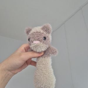 May include: A handmade plush toy in the shape of a small animal, possibly a mouse or a rat. The toy is primarily light brown with a darker brown head and small, black eyes. It has a pink nose and a fluffy texture.