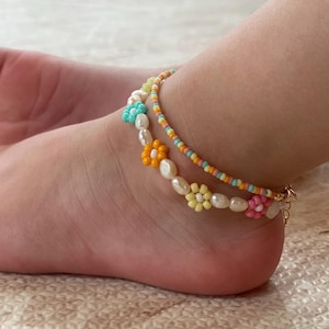 May include: Two beaded anklets. One features small, colorful beads in shades of orange, yellow, and blue. The other anklet has white pearl-like beads and flower-shaped beads in various colors, including yellow, orange, pink, and blue.
