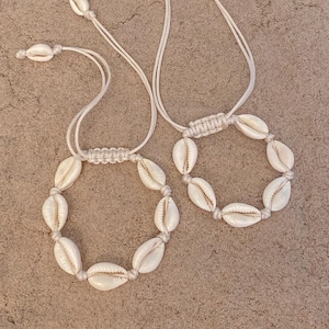 May include: Two seashell necklaces with white cord and adjustable closures. Each necklace features a circle of cream-colored seashells, connected by knotted cord. The necklaces are displayed on a sandy surface.