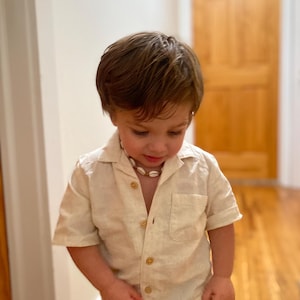 May include: A light beige linen shirt with short sleeves and a collar, paired with light blue denim jeans. The shirt has visible buttons and a chest pocket. A shell necklace is worn around the neck. The outfit is suitable for children.