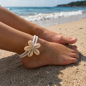 May include: A white anklet with a flower design made of seashells. The anklet is worn on a foot on a sandy beach with the ocean in the background. The anklet is made of white string and the flower is made of beige seashells.