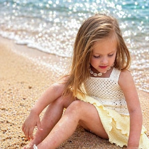 Könnte beinhalten: Ein junges Kind trägt ein cremefarbenes Häkelkleid und Muschelschmuck an einem Sandstrand. Das Kleid hat einen quadratischen Ausschnitt und einen gerüschten Rock. Eine Muschelkette und ein Fußkettchen vervollständigen den Strand-Look.