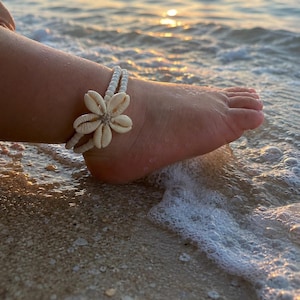 May include: A white beaded anklet with a flower made of seashells is worn on an ankle. The ankle is in the ocean with the sun setting in the background. The anklet is made of small white beads and seashells.