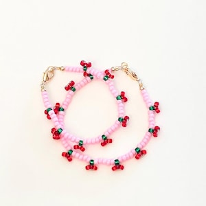 May include: Two beaded necklaces with a cherry design. The necklaces are made of pink beads with red and green cherry accents. The necklaces have gold-colored clasps.