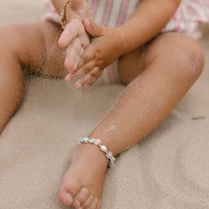 Peut inclure: Gros plan sur une cheville d'enfant ornée d'un bracelet délicat avec des perles et des fleurs aux couleurs pastel. L'enfant est assis sur une surface sablonneuse, les mains jouant avec le sable. Un deuxième bracelet est visible au poignet.