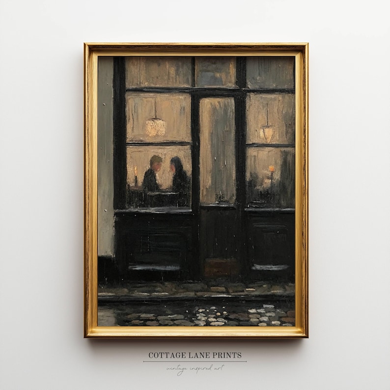 May include: A framed oil painting print depicting a dark storefront at night with two figures sitting inside by warm light. The scene features a cobblestone street in the foreground, large window panes, and a gold-coloured frame. Text reads COTTAGE LANE PRINTS.