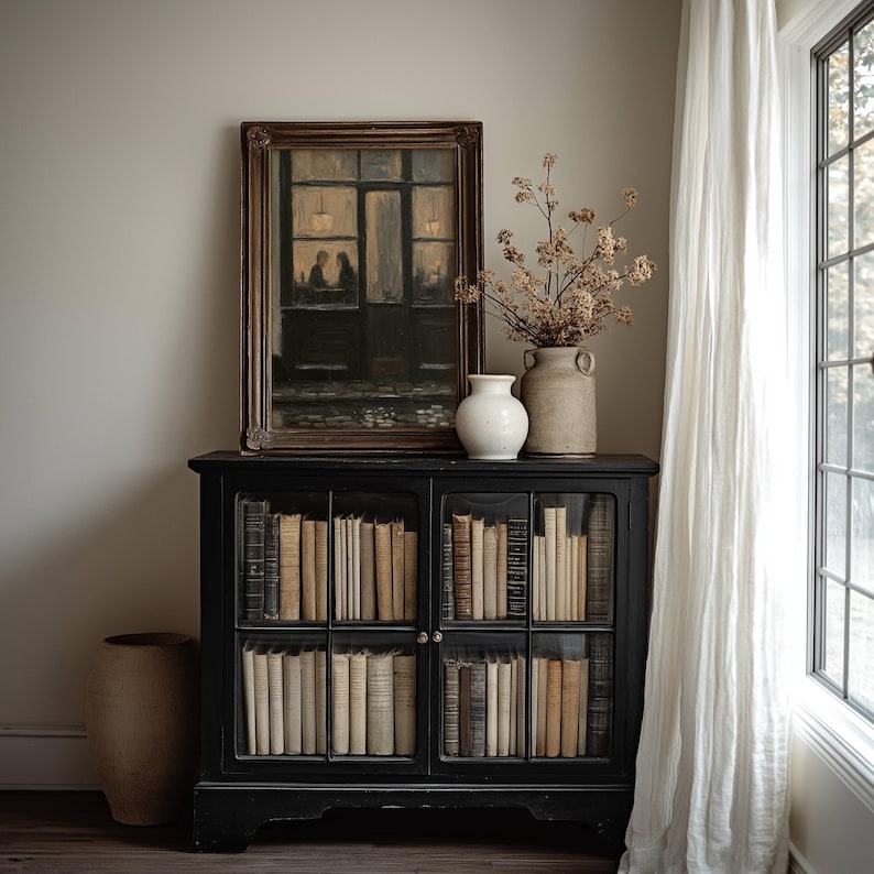 May include: A black wooden cabinet with glass doors filled with rows of books. On top sits a framed painting of a cafe scene, a white ceramic vase, and a tan ceramic vase containing dried branches. A large tan floor vase stands to the left.