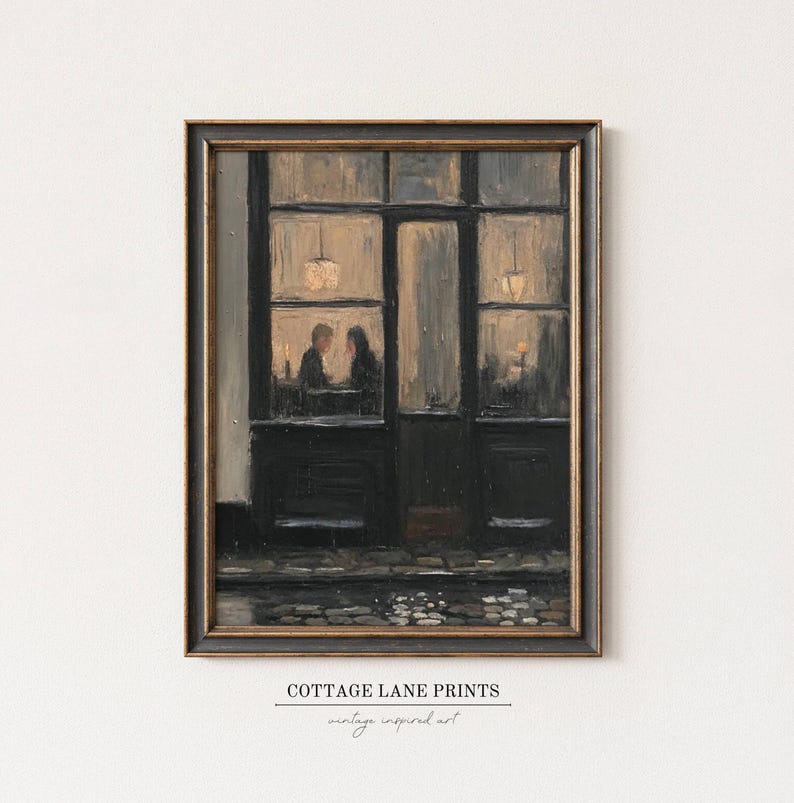 May include: A framed oil painting print depicting a dark storefront at night with two figures seated inside under warm glowing lights. The scene features a cobblestone street in the foreground and the text COTTAGE LANE PRINTS at the bottom.