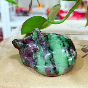 May include: A polished stone carving of a sleeping cat, crafted from zoisite with prominent green and black mineral inclusions and vibrant magenta ruby patches. The figurine features a smooth, rounded form with defined ears and a curled tail.