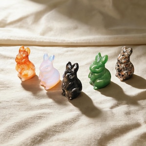 May include: Five small, carved rabbit figurines in a row. The rabbits are in shades of orange, opalescent white, black, green, and brown. The figurines are sitting on a cream-colored fabric.