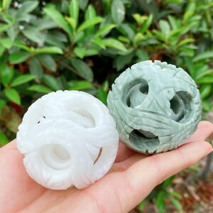 May include: Two carved stone spheres. One is white, the other is a pale green. Both spheres have intricate, openwork designs. The white sphere has a swirling pattern, while the green sphere has a face-like design. The background is blurred green foliage.