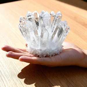 Pode incluir: Um aglomerado de cristais de quartzo transparente, com múltiplas pontas, segurado em uma mão. Os cristais estão dispostos em forma circular, com uma base de cristais menores e granulares. O quartzo é translúcido.