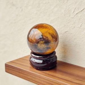 May include: A polished tiger's eye sphere, showcasing bands of golden brown and amber hues. The gemstone sits atop a dark, glossy wooden stand. The sphere is displayed on a wooden shelf against a neutral background.