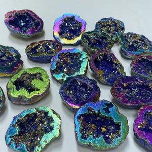 May include: A collection of iridescent geode clusters. The geodes have a dark, crystalline interior and are surrounded by a colorful, metallic-like exterior with hues of blue, green, and purple. The geodes are irregularly shaped.