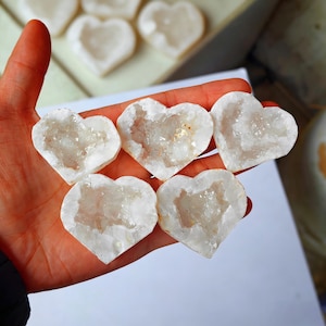 Könnte beinhalten: Eine Sammlung weißer, herzförmiger Geodensteine, jeweils mit kristallinem Innenraum. Die Geoden werden in einer Hand gehalten, weitere sind im Hintergrund zu sehen. Die Steine haben ein natürliches, organisches Aussehen.
