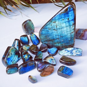 Könnte beinhalten: Eine Sammlung polierter Labradoritsteine in verschiedenen Formen und Größen. Die Steine zeigen irisierende blaue, grüne und goldene Reflexe. Ein großes, dreieckiges Stück ist der Mittelpunkt, umgeben von kleineren Trommelsteinen.