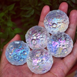 May include: Five iridescent glass spheres with a textured surface resembling the moon's craters. The spheres reflect a spectrum of colors, including blue, purple, and yellow. The spheres are held in a hand against a backdrop of green foliage.