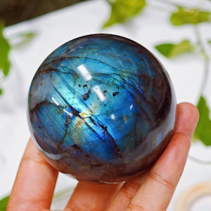 以下が含まれることがあります： 磨かれたラブラドライトの球体で、魅惑的な色の遊びを披露しています。球体の表面は、虹色の青、緑、金色のヒントを表示します。宝石は手で持たれており、そのサイズと自然の美しさを強調しています。装飾品です。