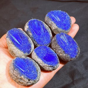 Può includere: Sei pietre ovali con un sorprendente centro blu, simile a cristalli. Le pietre hanno un esterno ruvido e grigio. I cristalli blu sembrano irradiare dal centro di ogni pietra. Le pietre sono tenute in una mano.
