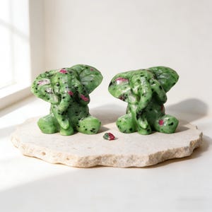 May include: Two green stone elephant figurines with red accents sit on a light-colored, textured stone. The elephants have detailed trunks, ears, and bodies. A small piece of the same stone is in front of them.