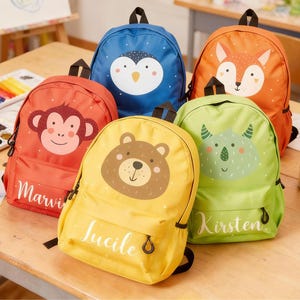 Peut inclure: Cinq sacs à dos colorés avec des visages d'animaux : un singe, un ours, un pingouin, un renard et un rhinocéros. Chaque sac à dos a une couleur différente et un nom imprimé sur le devant. Les sacs à dos sont sur une table en bois.