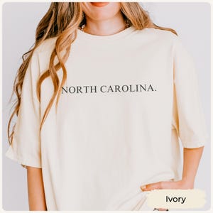 May include: Ivory-coloured t-shirt with the text "NORTH CAROLINA." printed in black. The shirt has short sleeves and a relaxed fit. The shirt is made of a soft, comfortable material.