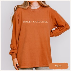 May include: A long-sleeved, crew neck shirt in a warm yam colour. The shirt features the text "NORTH CAROLINA." in white, centered on the chest. The sleeves are long and the fabric appears soft. The shirt is a casual style.