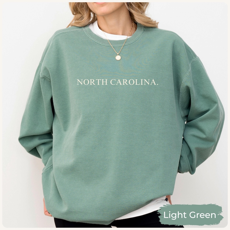 May include: Light green sweatshirt with the text "NORTH CAROLINA." in white. The sweatshirt has long sleeves and a crew neck. The bottom right corner of the image has the text "Light Green."