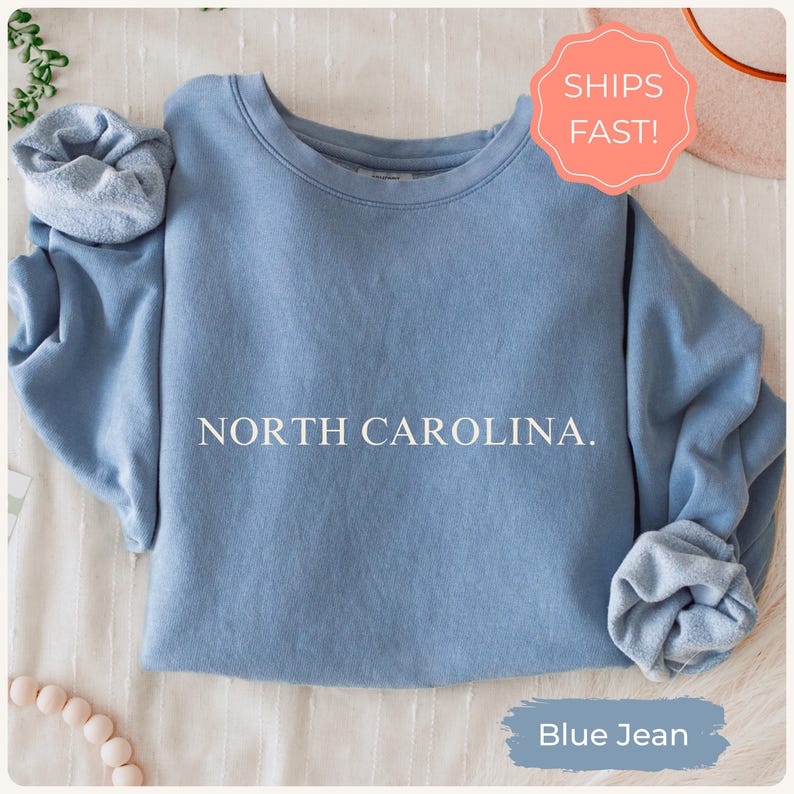 May include: A blue jean-coloured sweatshirt with the text "NORTH CAROLINA." printed in white. The sleeves are rolled up. A sticker that says "SHIPS FAST!" is in the upper right corner. The words "Blue Jean" are at the bottom.