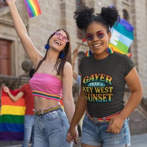 May include: Two people holding hands, celebrating Pride. One wears a black t-shirt with the text "Gayer than a Key West Sunset." The other wears a pink top and jeans. Both have rainbow accessories and flags.