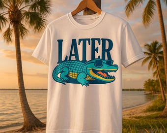 May include: A white short-sleeve t-shirt featuring a graphic of a teal alligator wearing black sunglasses. Above the alligator, the word LATER is printed in large, bold, dark blue capital letters. The design includes yellow accents on the alligator.