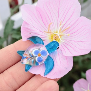May include: A miniature glass turtle ornament with a clear body containing colourful flecks and bright blue flippers and head. The turtle is placed on a pink flower with a yellow centre. The ornament is held in a person's hand.