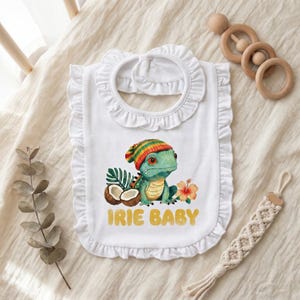 May include: White baby bib with a ruffled edge and a neck tie. The bib features a colorful illustration of a lizard wearing a hat, with the words "IRIE BABY" in gold lettering. The design includes coconuts, tropical leaves, and a flower.