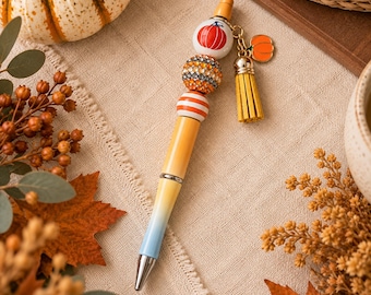 May include: A ballpoint pen featuring a yellow to light blue gradient barrel, topped with three decorative beads including a white pumpkin bead, a rhinestone-encrusted orange bead, and an orange-and-white striped bead, plus a gold tassel charm.