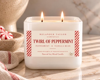 May include: A white square soy blend candle with two lit wicks. The label reads: MELANDER TAYLOR COMPOSES TWIRL OF PEPPERMINT PEPPERMINT • VANILLA BEAN HAND-POURED IN MASSACHUSETTS Natural Soy Blend Candle. The label has red striped borders.