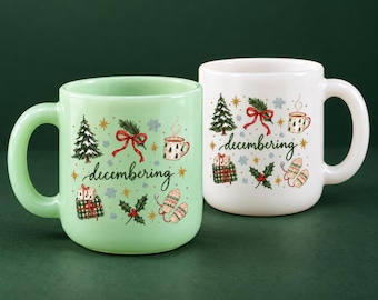 May include: A light green mug and a white mug, both featuring the word "decembering" in cursive script surrounded by illustrations of Christmas trees, wrapped gifts, red bows, holly leaves, steaming mugs, and patterned mittens.