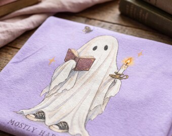 May include: A light purple t-shirt featuring a graphic of a white ghost wearing black sneakers, reading a brown book, and holding a lit candle. A small butterfly is above the ghost, and the text MOSTLY HARMLESS is printed at the bottom in black.