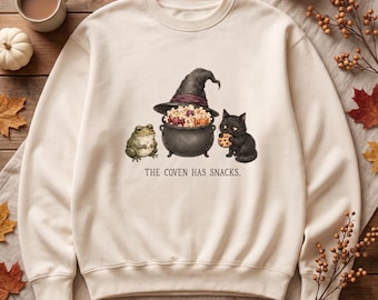 May include: A cream-colored crewneck sweatshirt featuring a graphic of a green frog, a black cauldron filled with popcorn, and a black cat eating a chocolate chip cookie. Below the illustration, the text reads, THE COVEN HAS SNACKS.