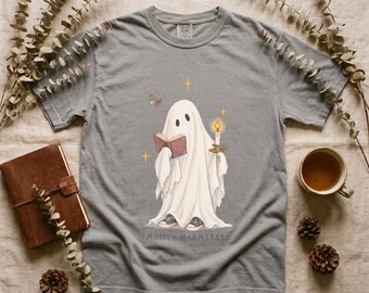 May include: A grey short-sleeve t-shirt featuring a graphic of a white ghost wearing sneakers, reading a book, and holding a lit candle. The text "MOSTLY HARMLESS" is printed below the ghost, surrounded by small star and butterfly illustrations.