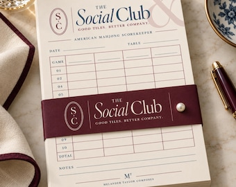 May include: A cream-colored American Mahjong scorekeeper pad titled The Social Club, Good Tiles, Better Company. It features a dark maroon paper band with a pearl accent, a grid for game scores, and the text Melander Taylor Composes at the bottom.