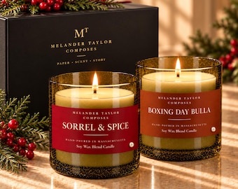 May include: Two soy wax blend candles in amber glass jars with lit wicks. One candle has a red label reading Sorrel & Spice, the other an orange label reading Boxing Day Bulla. Both feature the Melander Taylor Composes brand name.